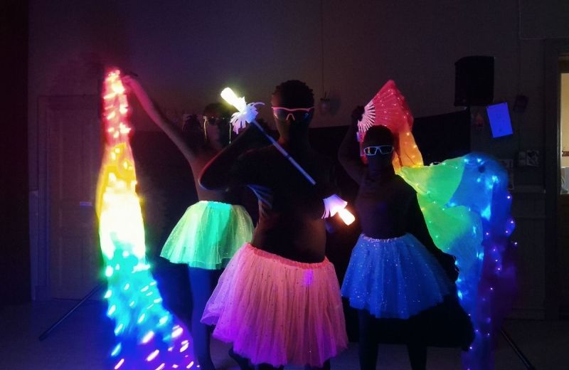 Tre personer i lysande tyllkjolar dansar med färgglada LED-slöjor och lysstavar i ett mörkt rum.