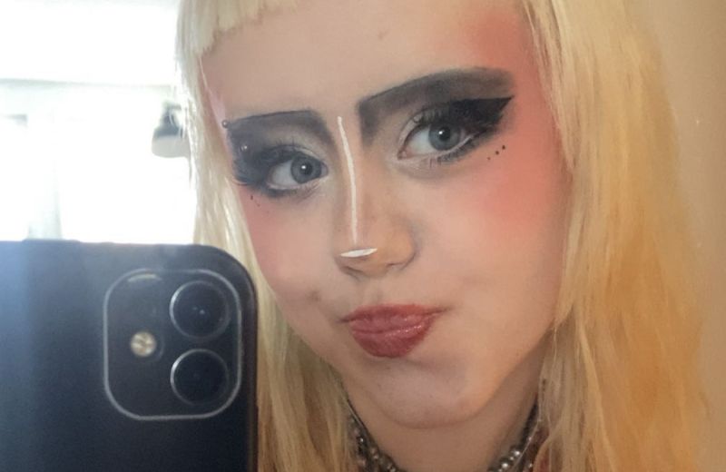 En närbild på en Doris med blont hår och kreativ sminkning som tar en selfie i en spegel.