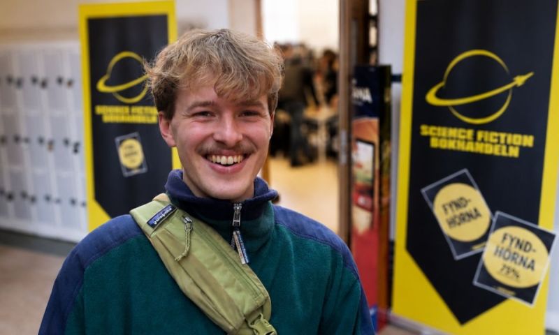 En leende person i grön fleece och axelväska står framför skyltar för Science Fiction Bokhandeln. Bakgrunden är lätt suddig med entré och besökare bakom.