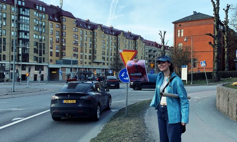 Ung tjej i blå jacka står vid en trafikerad väg och visar stolt upp en affisch med text.