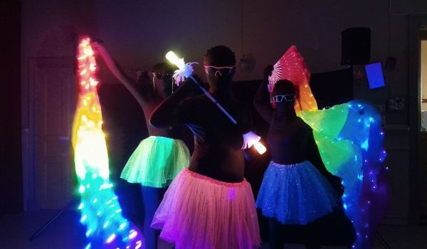 Tre personer i lysande tyllkjolar dansar med färgglada LED-slöjor och lysstavar i ett mörkt rum.