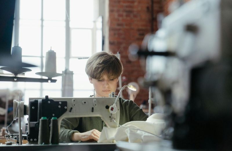 En person syr i vitt tyg med en industrisymaskin i en lokal med tegelväggar.