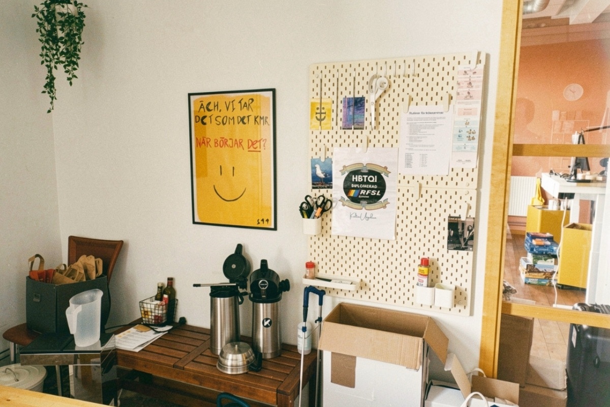 En kontorsvägg med gul affisch, verktygstavla och kaffetermosar på en träbänk.