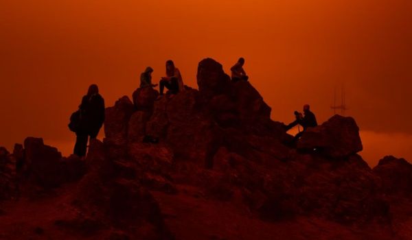 Silhuetter av personer som sitter på klippor mot en starkt orange, dimmig himmel.