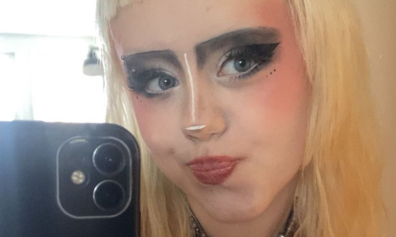 En närbild på en Doris med blont hår och kreativ sminkning som tar en selfie i en spegel.