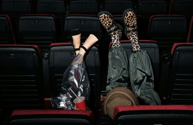 Två unga personer med uppfällda ben i en biosalong, ena med leopard skor, andra med tatuerat ben.