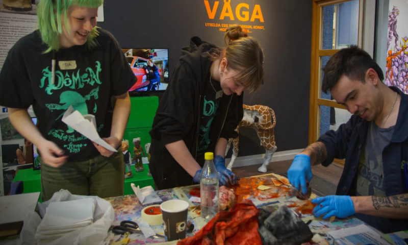 Tre personer skapar tillsammans vid ett stökigt arbetsbord fullt av färg och verktyg