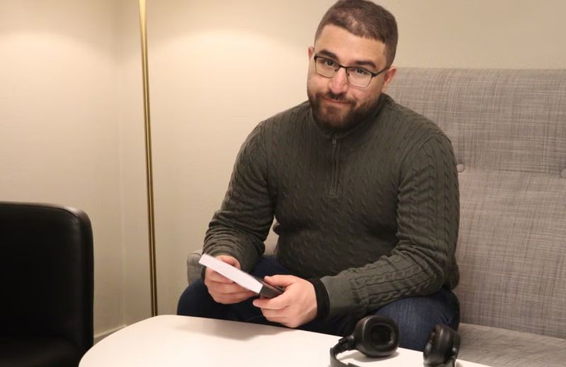 Albaraa Abdulrahman sitter i en soffa vid ett runt vitt bord och håller i sin bok. Han har glasögon, mörkgrön stickad tröja och jeans. På bordet ligger ett par hörlurar.