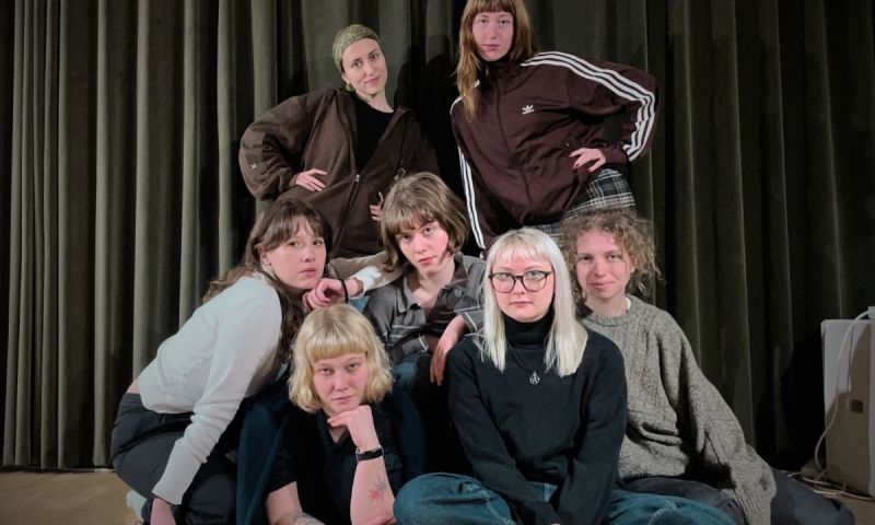 Nära gruppbild på sju unga vuxna i olika stilar, mot mörk drakplagg.