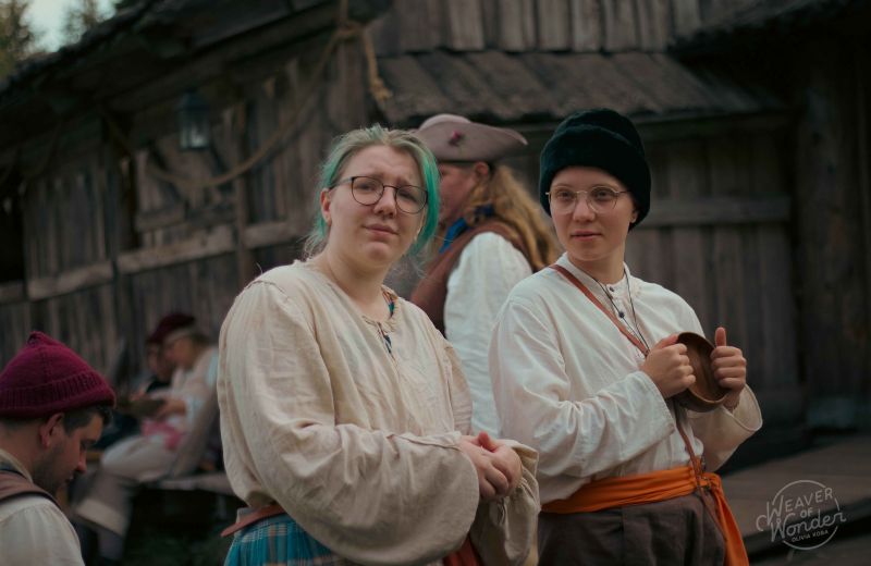 Deltagare i tidstypiska dräkter och glasögon blickar framåt under ett lajv i en historisk trämiljö.