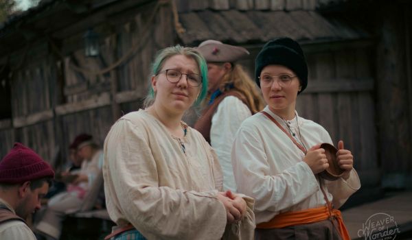 Deltagare i tidstypiska dräkter och glasögon blickar framåt under ett lajv i en historisk trämiljö.