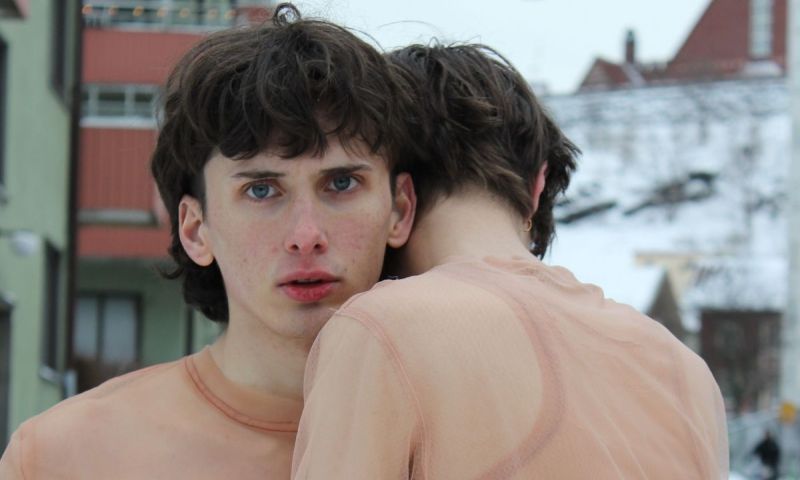 Två personer i hudfärgade nätkläder kramas utomhus i en stadsmiljö.