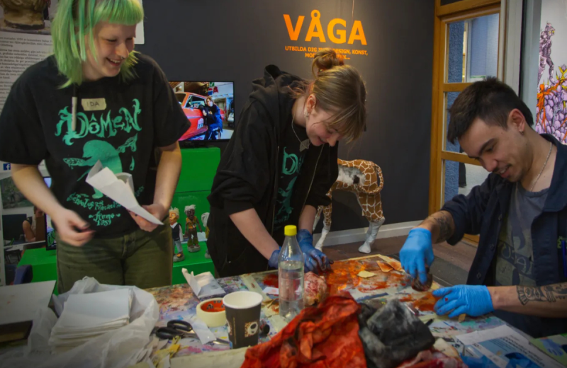 Tre personer skapar tillsammans vid ett stökigt arbetsbord fullt av färg och verktyg