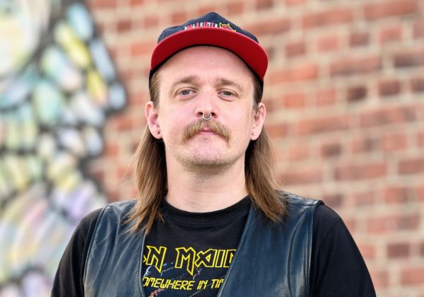 Porträtt av Robin Havbring med axellångt brunt hår, mustasch och näsring. Han bär en rödsvart keps, en svart t-shirt med Iron Maiden-tryck och en skinnväst framför en graffitivägg.
