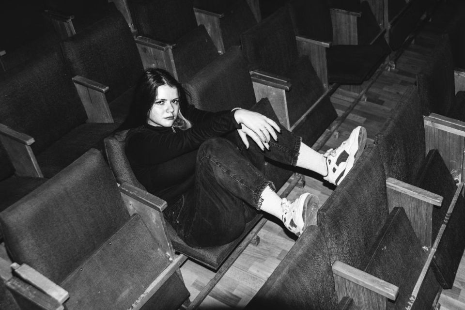 Svartvit bild på en ung kvinna som sitter i en tom biosalong och vilar fötterna på sätet framför.