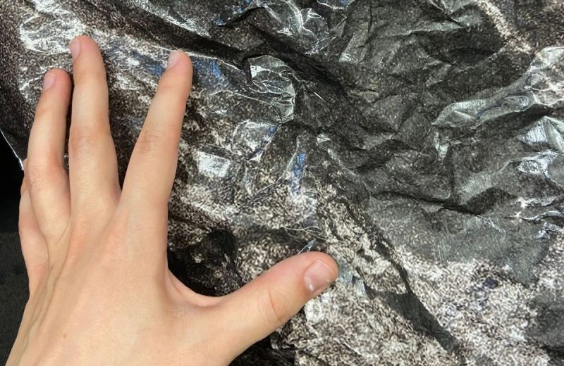 En hand vilar mot ett skrynkligt, silvrigt och mörkt material med en grov textur, som ska likna en sten.