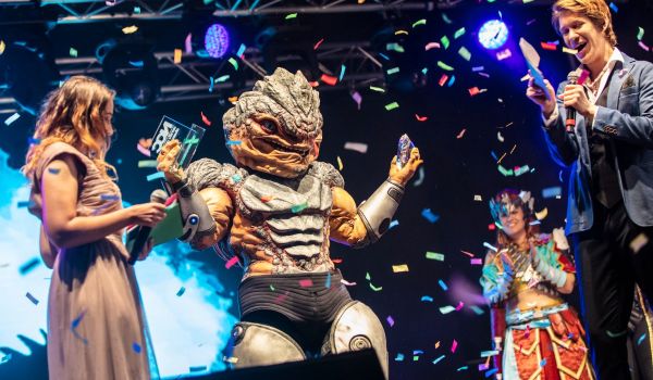 En cosplayer i detaljrik reptilliknande rustning håller upp två priser på en scen medan färgglad konfetti regnar ner och personer runt omkring firar.