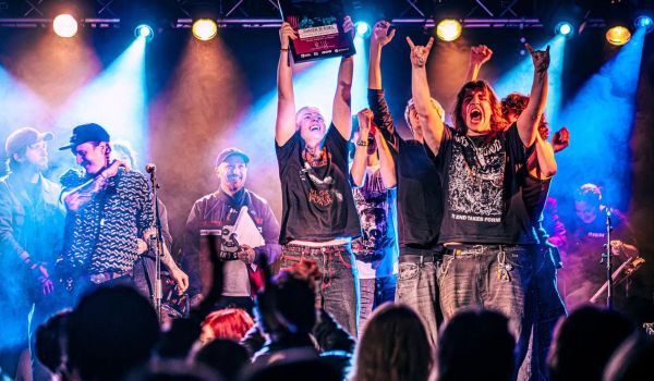 Metalbandet Awaken in Ashes jublar på scen och håller upp ett diplom inför en publik i färgstarkt scenljus.