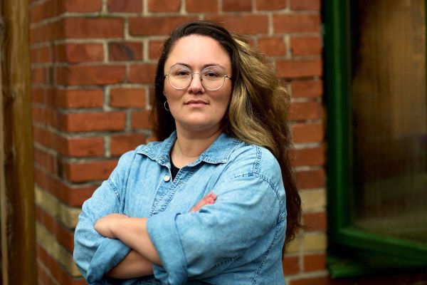 Porträtt av Yllka Sheqiri med långt brunt hår och glasögon. Hon står med armarna i kors och bär en blå jeansskjorta framför en röd tegelvägg med en grön fönsterkarm.