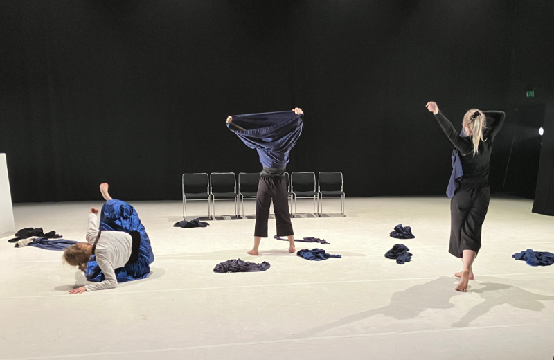 Dansare i rörelse på en scen med blå tyger, där en person drar en tröja över huvudet i ett uttrycksfullt ögonblick.