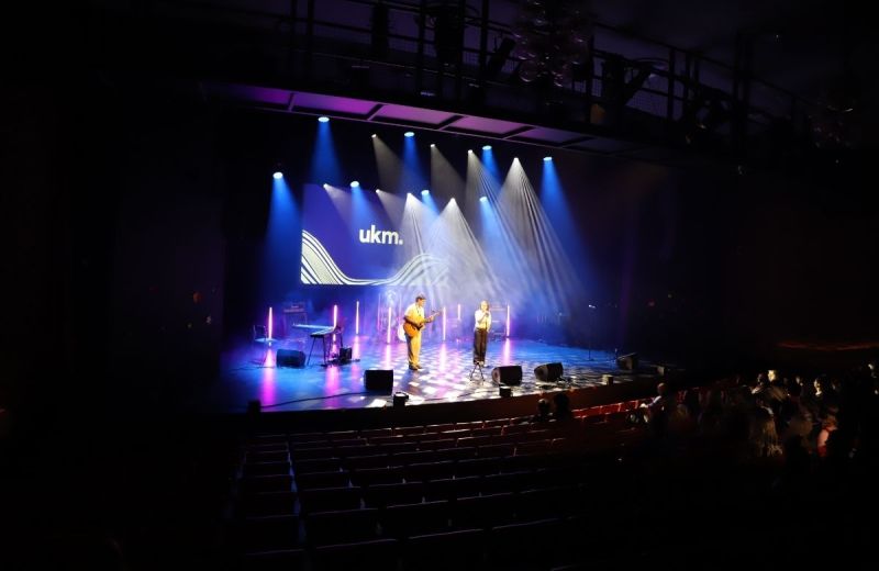 Två personer uppträder på en stor scen med strålkastare och UKM-logotypen på en skärm i bakgrunden.