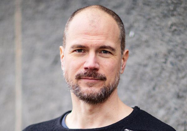 Porträtt av Erik Lundin med kortklippt mörkt hår och skägg. Han ser in i kameran med en lugn blick och bär en svart tröja framför en vägg.