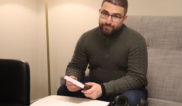 Albaraa Abdulrahman sitter i en soffa vid ett runt vitt bord och håller i sin bok. Han har glasögon, mörkgrön stickad tröja och jeans. På bordet ligger ett par hörlurar.