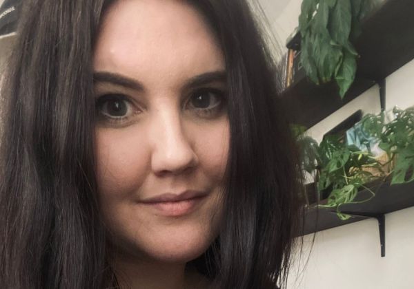 Ett närporträtt av Elina Andersson med långt, mörkt vågigt hår. Hon ler mjukt mot kameran i en miljö med gröna växter på hyllor i bakgrunden.