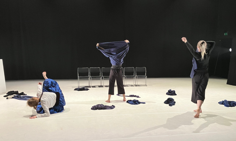 Dansare i rörelse på en scen med blå tyger, där en person drar en tröja över huvudet i ett uttrycksfullt ögonblick.