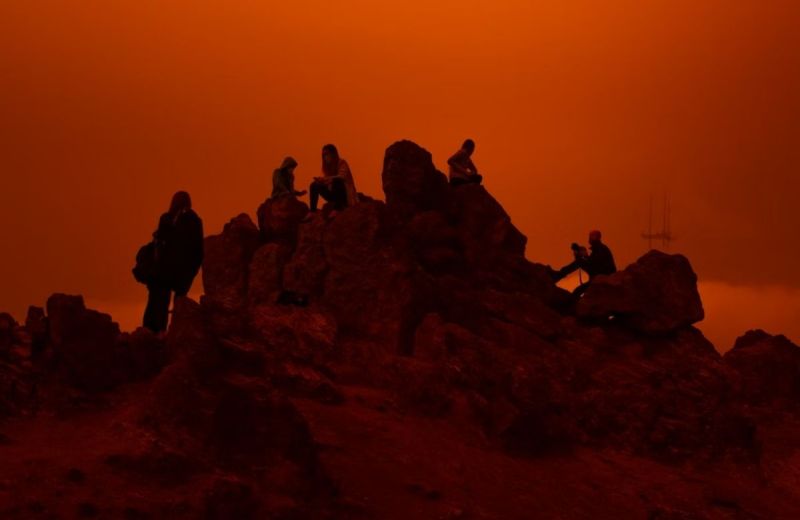 Silhuetter av personer som sitter på klippor mot en starkt orange, dimmig himmel.