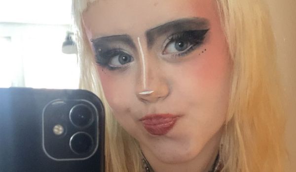 En närbild på en Doris med blont hår och kreativ sminkning som tar en selfie i en spegel.