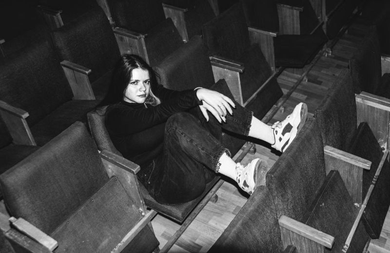 Svartvit bild på en ung kvinna som sitter i en tom biosalong och vilar fötterna på sätet framför.