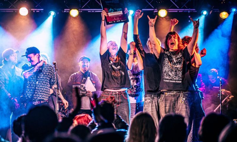 Metalbandet Awaken in Ashes jublar på scen och håller upp ett diplom inför en publik i färgstarkt scenljus.