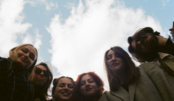 Sex ungdomar tittar ner i kameran underifrån mot en ljusblå himmel med vita moln.