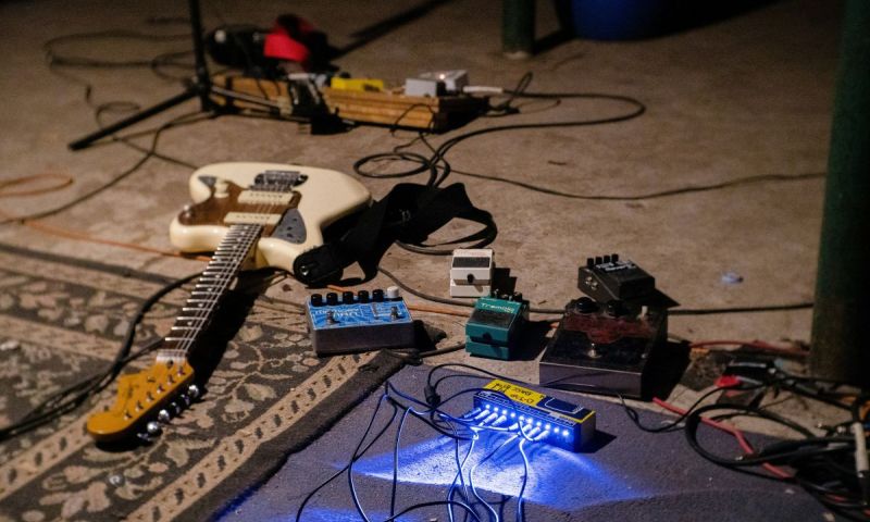 En vit elgitarr ligger på golvet omgiven av effektpedaler och sladdar i en replokal.
