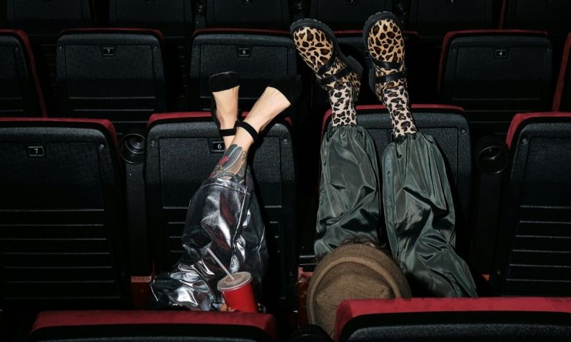 Två unga personer med uppfällda ben i en biosalong, ena med leopard skor, andra med tatuerat ben.