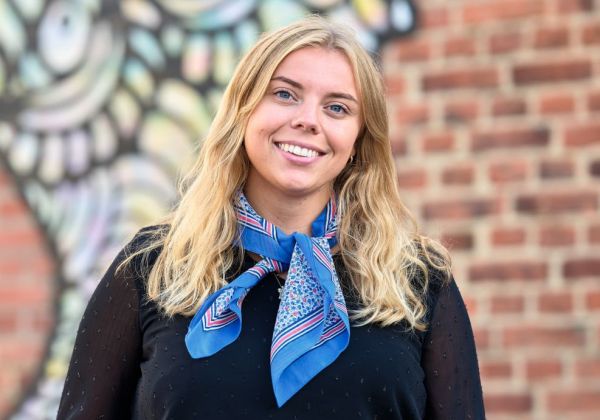 Porträtt av en leende Alva Pettersson med långt blont hår. Hon bär en svart blus och en blåmönstrad scarfs knuten runt halsen framför en tegelvägg med graffiti.