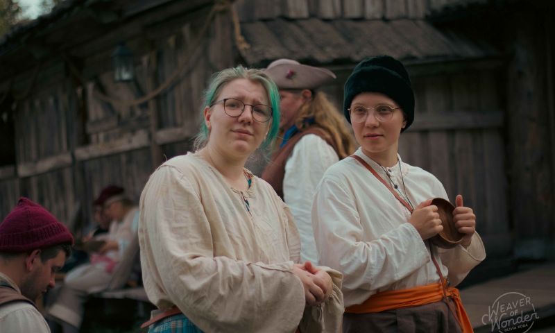 Deltagare i tidstypiska dräkter och glasögon blickar framåt under ett lajv i en historisk trämiljö.