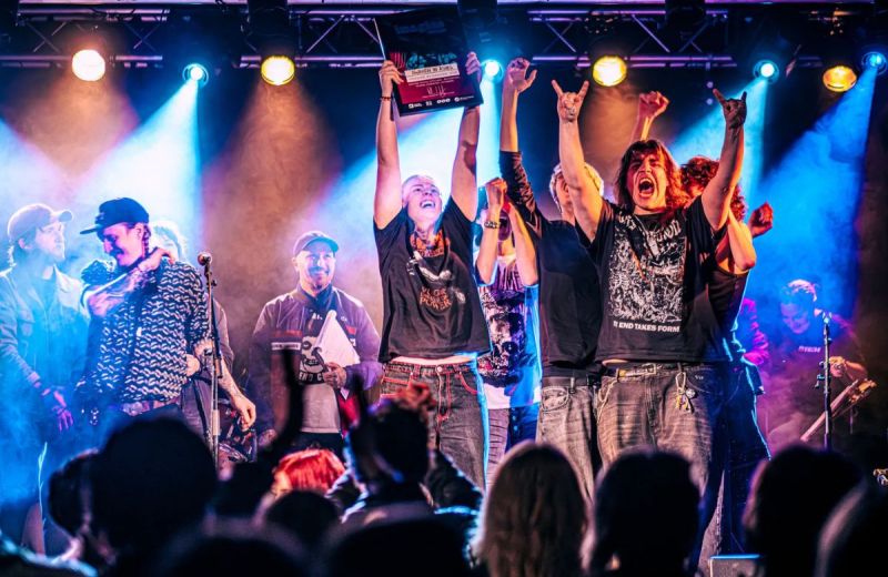 Metalbandet Awaken in Ashes jublar på scen och håller upp ett diplom inför en publik i färgstarkt scenljus.