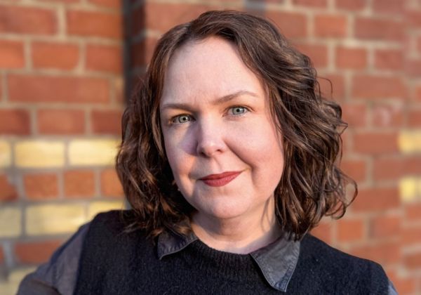 Ett porträtt av Matilda Klamas med axellångt vågigt brunt hår och rött läppstift. Hon bär en svart väst över en mörk tröja och står framför en röd tegelvägg.