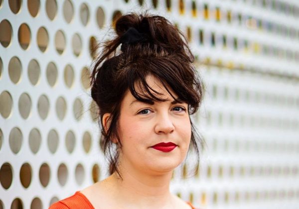 Porträtt av Elin Åström med mörkt hår uppsatt i en hög knut och rött läppstift. Hon bär en röd topp och står framför en vit vägg med ett runt mönster.