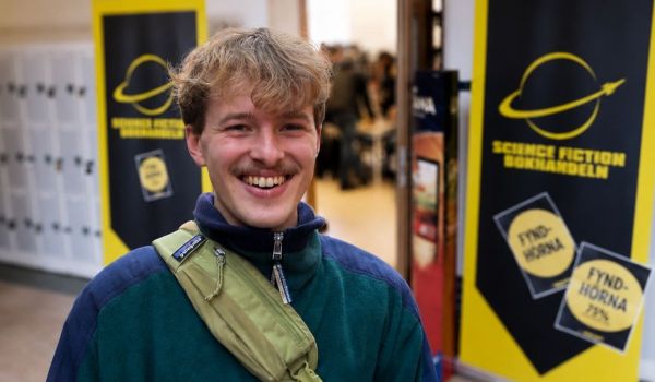 En leende person i grön fleece och axelväska står framför skyltar för Science Fiction Bokhandeln. Bakgrunden är lätt suddig med entré och besökare bakom.