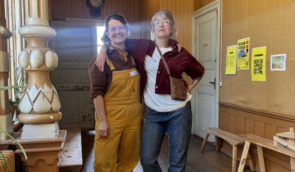 Två glada personer står omfamnade i en rustik trälokal med festivalaffischer och träbänkar.