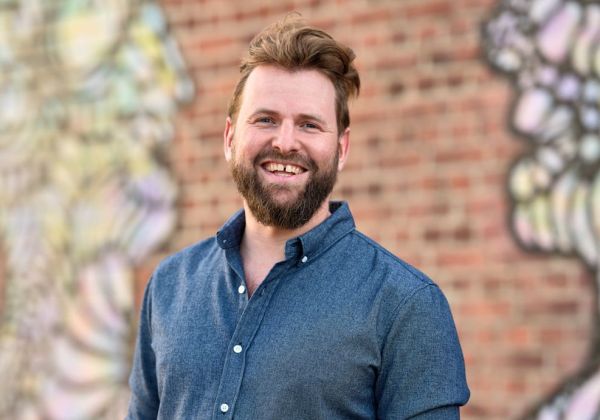 Porträtt av en leende Nils Lindenmo med kort brunt hår och skägg. Han bär en blå jeansskjorta framför en bakgrund med graffitikonst.