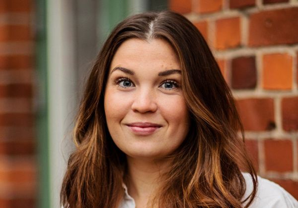 Porträtt av en leende Gabriella Ortiz med långt brunt hår. Hon bär en vit skjorta och står framför en röd tegelvägg.