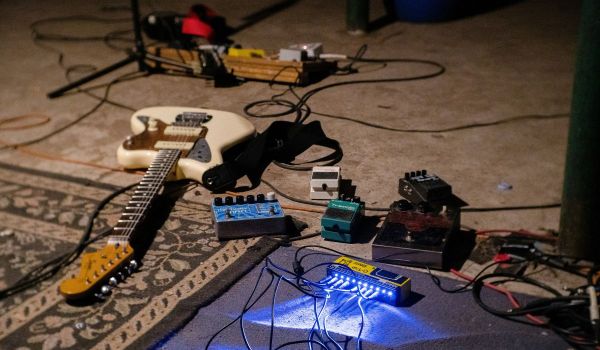 En vit elgitarr ligger på golvet omgiven av effektpedaler och sladdar i en replokal.