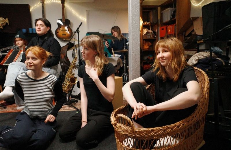 En grupp unga personer i en kreativ repetitionsmiljö med instrument som saxofon, gitarr och dragspel i bakgrunden. En person sitter i en stor flätad korg och ler.
