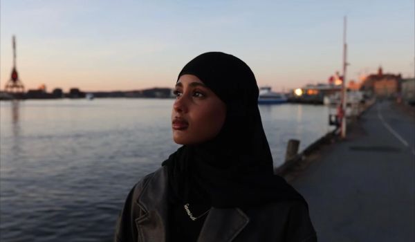 Porträtt i skymningsljus på Samira Hassan i slöja som står vid en kaj med stad och vatten i bakgrunden.