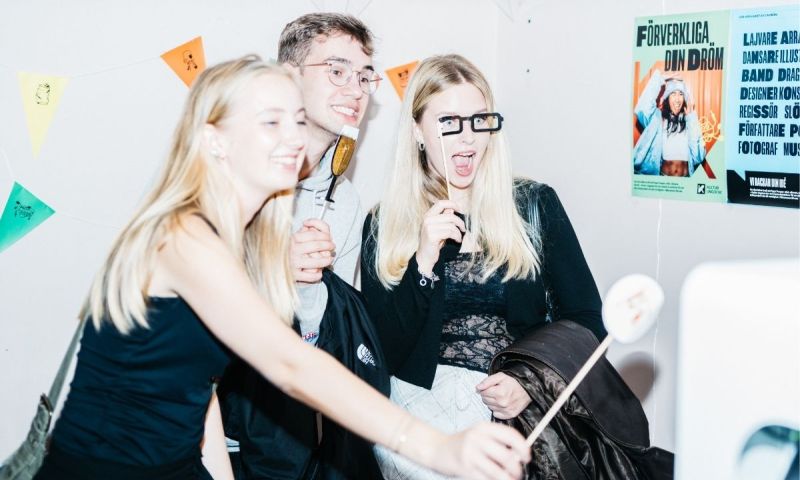 Tre glada ungdomar spexar med fotorekvisita framför en vit vägg med vimplar och affischer.