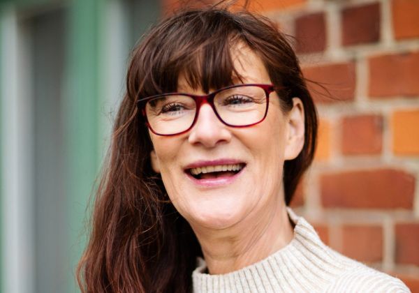 Ett porträtt av en leende Suzanne Johansson med runda röda glasögon och långt brunt hår. Hon bär en ljus polotröja och står framför en röd tegelvägg.
